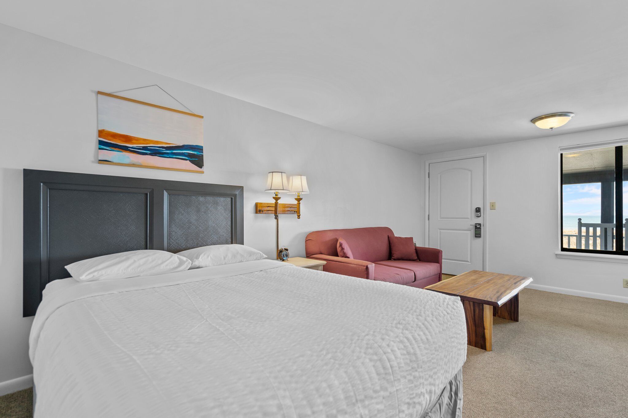A hotel room with a large bed, a desk, a chair, a lamp, and a painting on the wall.
