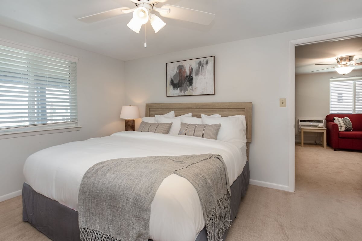 A bedroom with a bed, a ceiling fan, a bedside table with a lamp, a red sofa, and a painting on the wall.