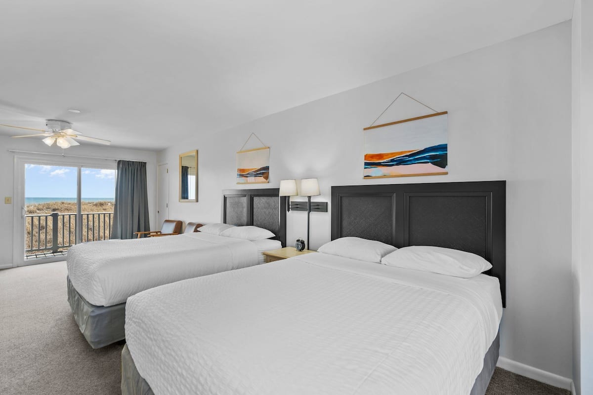 A hotel room with two beds, a ceiling fan, a balcony with a view, and artwork on the walls.