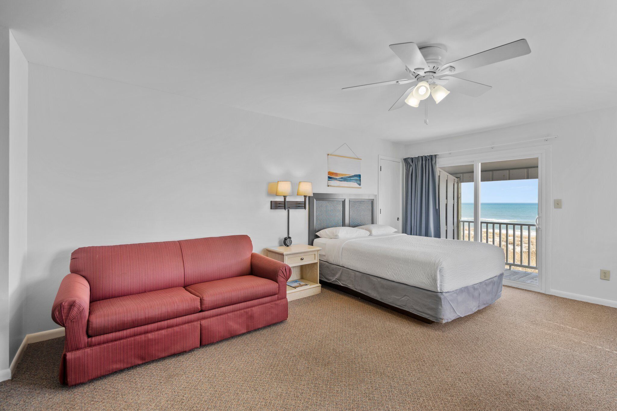 A hotel room with a red sofa, a bed, a ceiling fan, a nightstand with lamps, and a view of the beach through a sliding door.