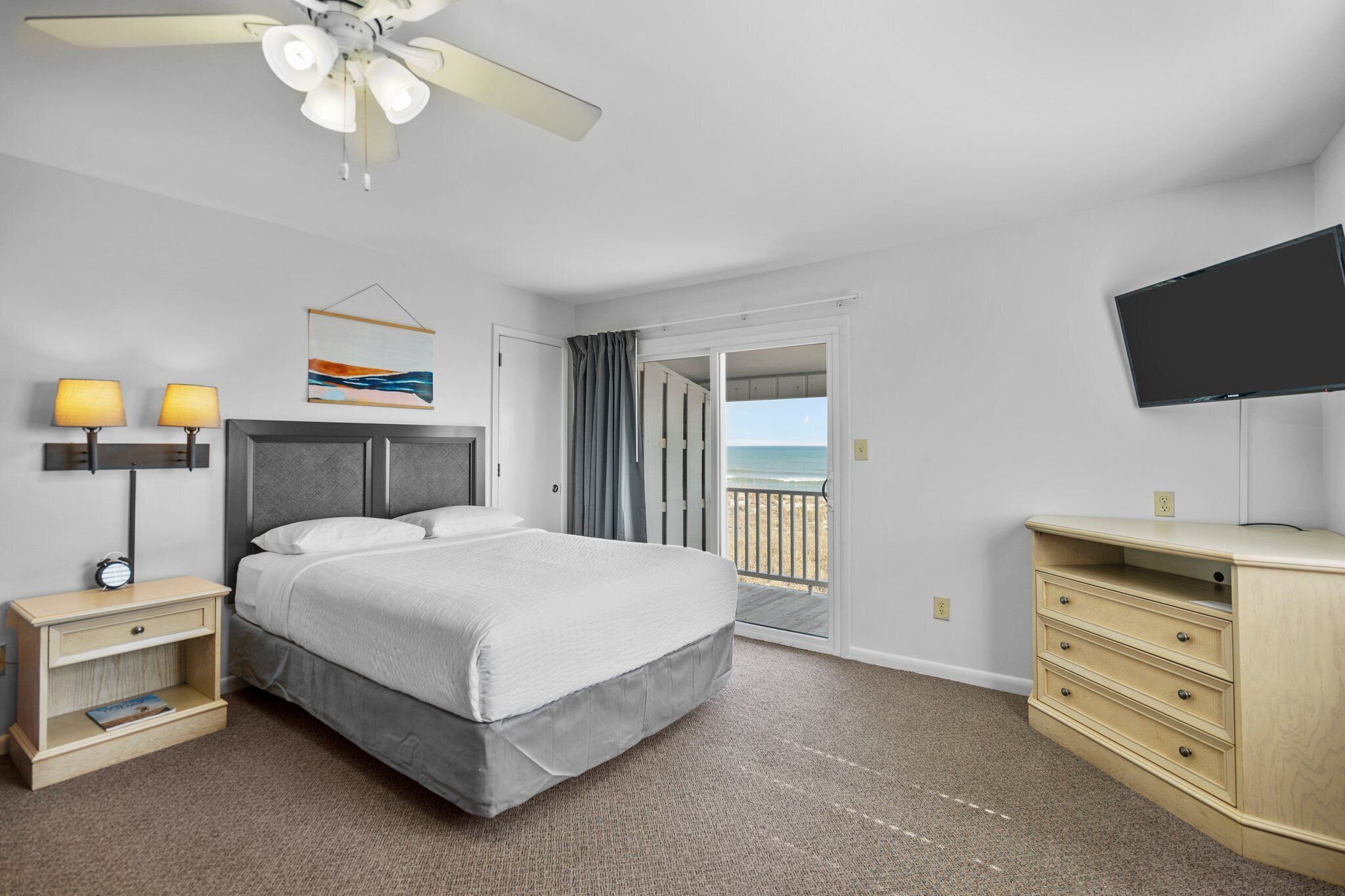 A hotel room with a bed, a nightstand, a dresser, a television, and a view of the beach.
