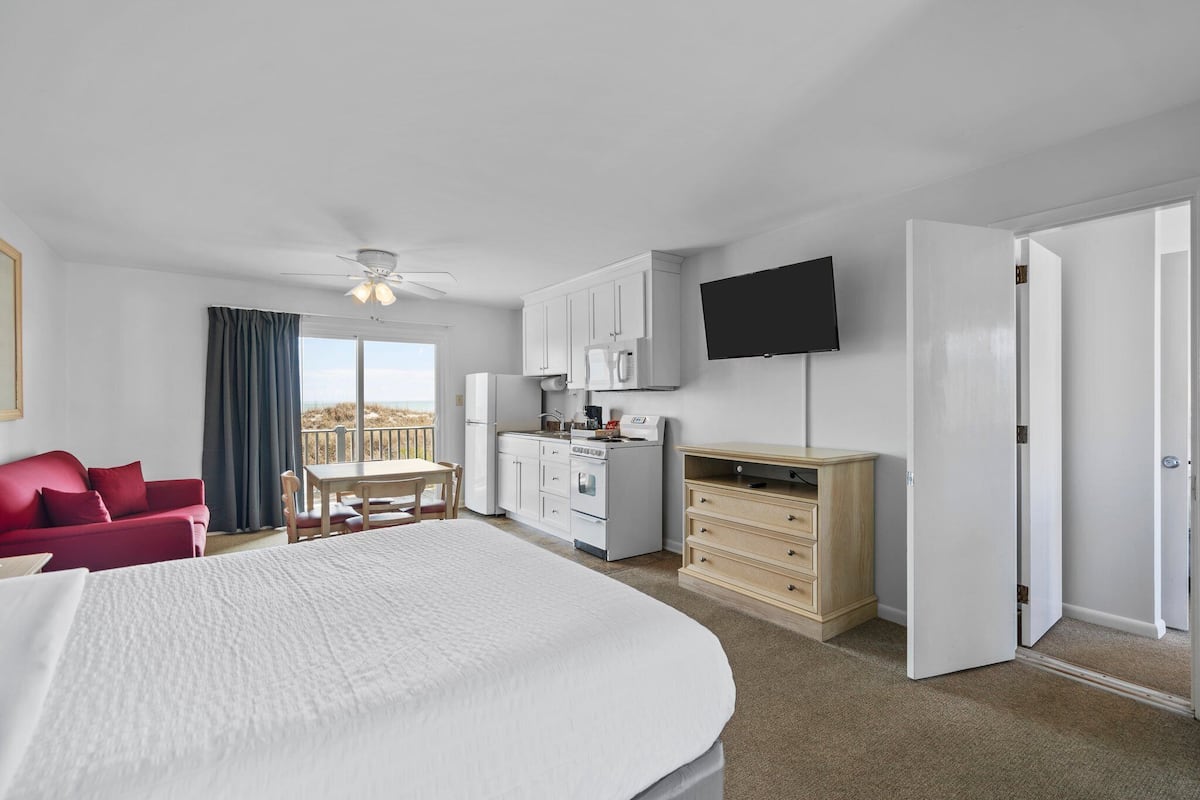 A hotel room with a large bed, a dining table, a kitchenette, a TV, and a balcony.