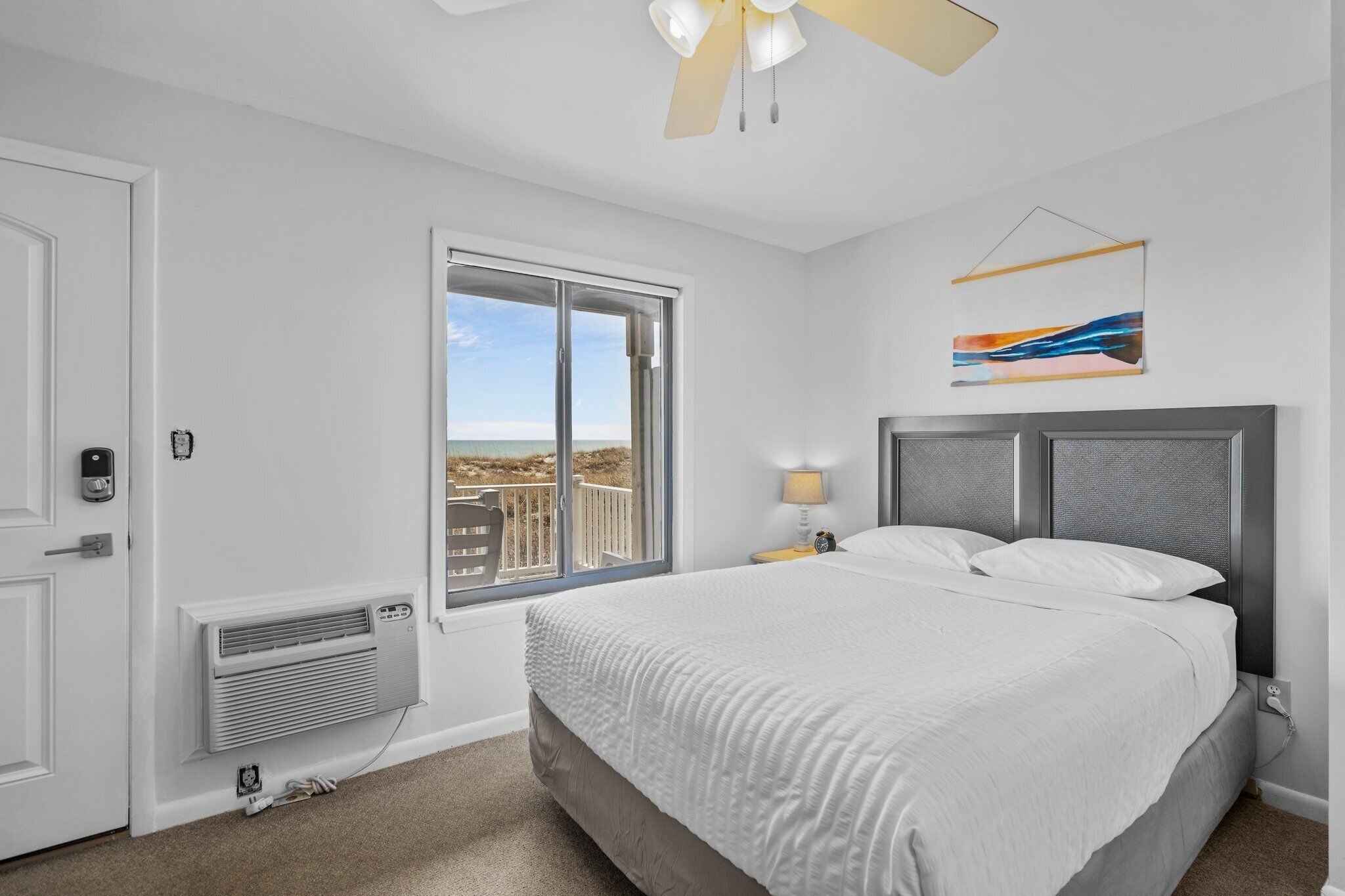A bedroom with a bed, a window with a view, a ceiling fan, and a wall-mounted artwork.
