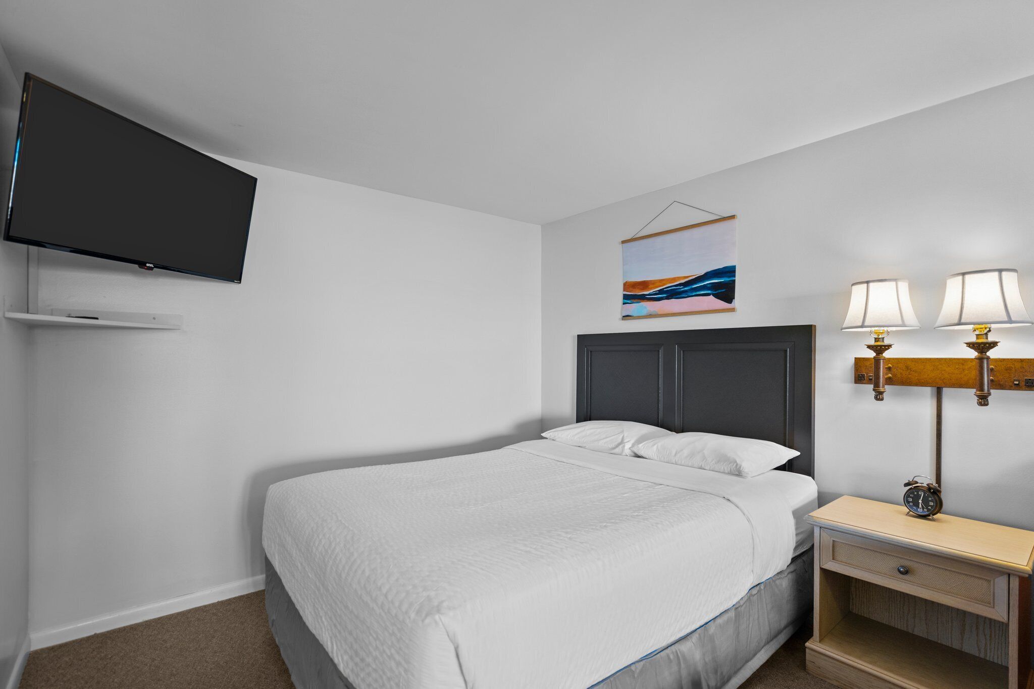 A hotel room with a bed, a TV mounted on the wall, a bedside table with lamps, and a painting on the wall.