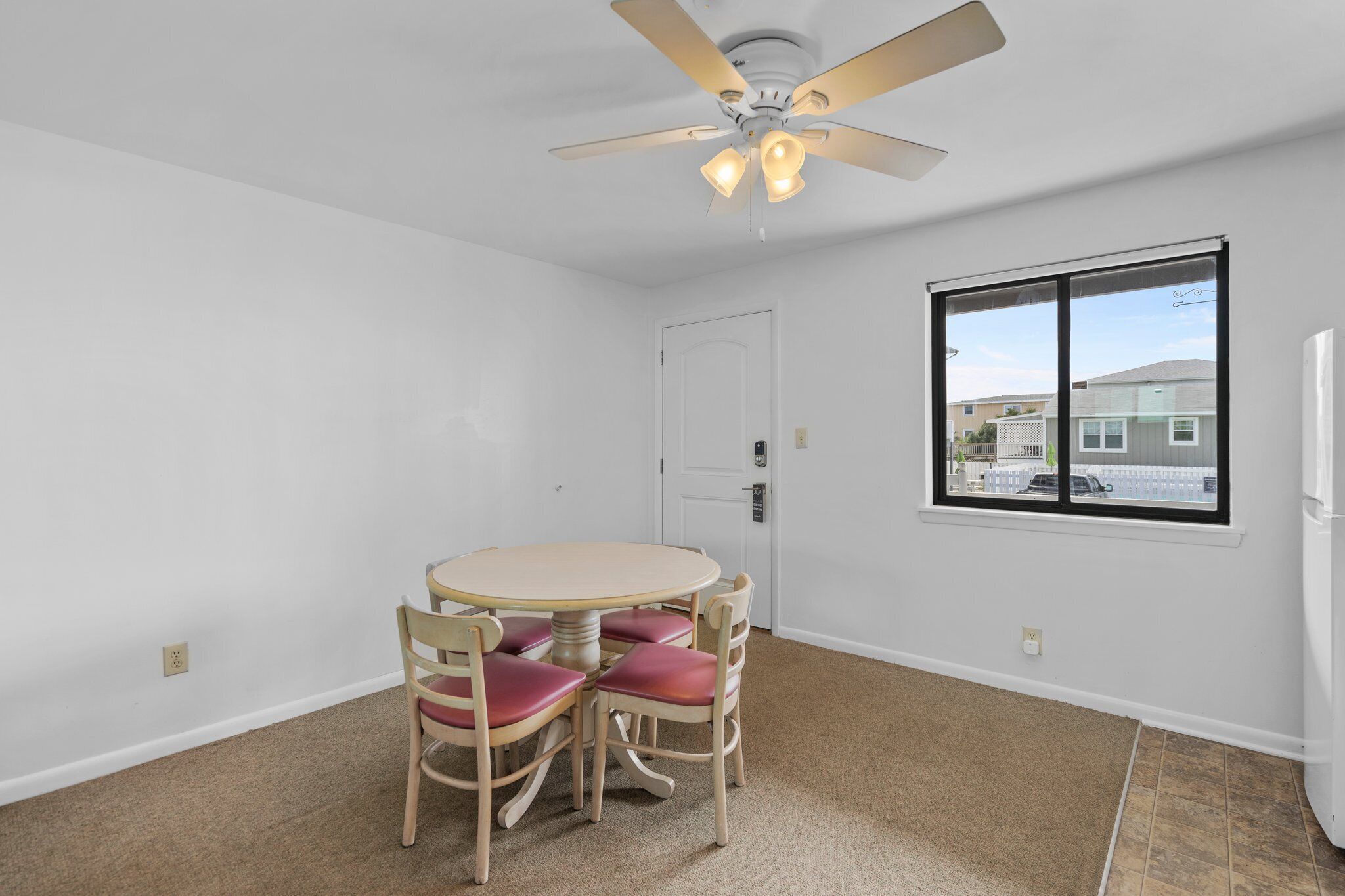 A room with a round table and four chairs, a ceiling fan, a window with a view of houses, and a door.