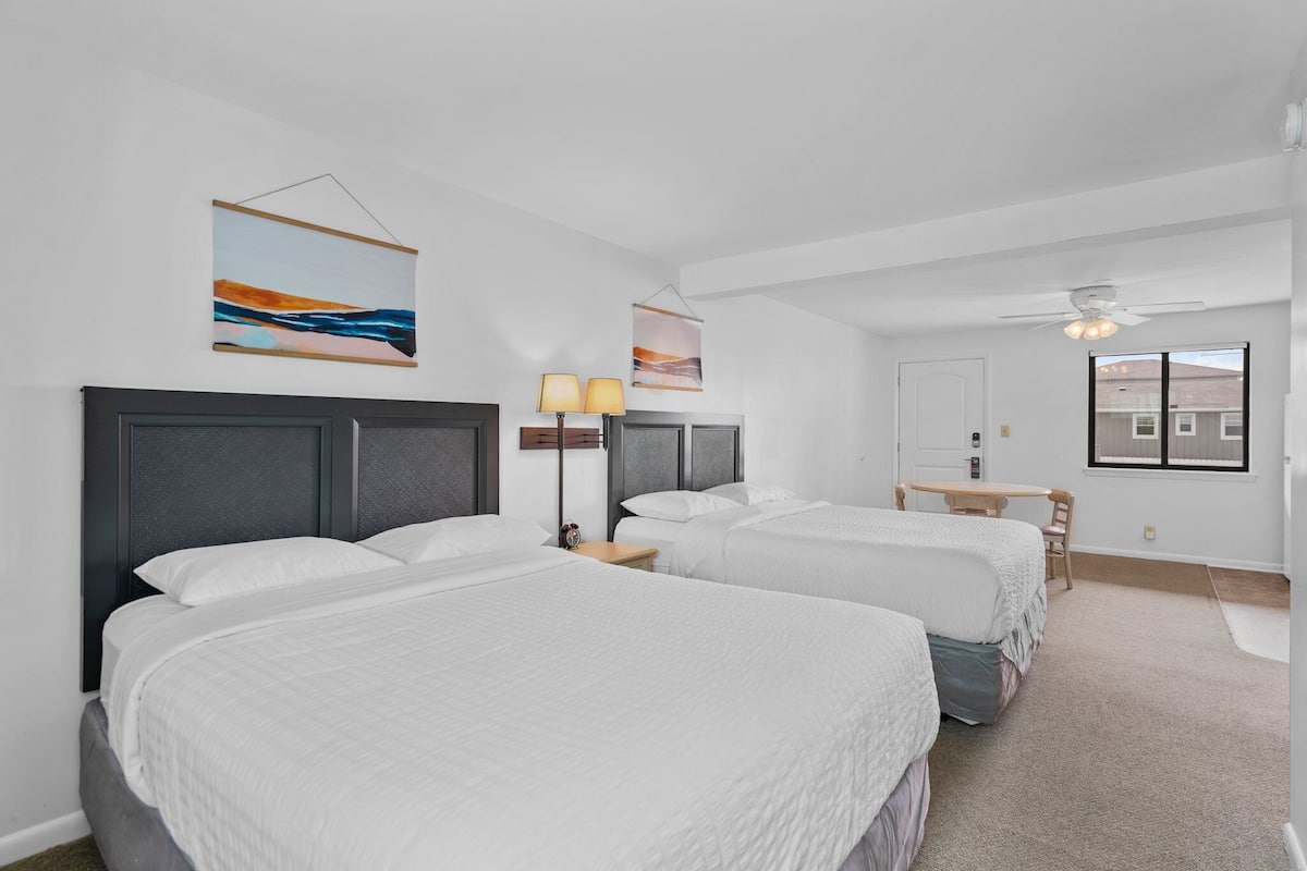 A hotel room with two beds, a desk, and a chair. There are two wall-mounted lamps and a ceiling fan.