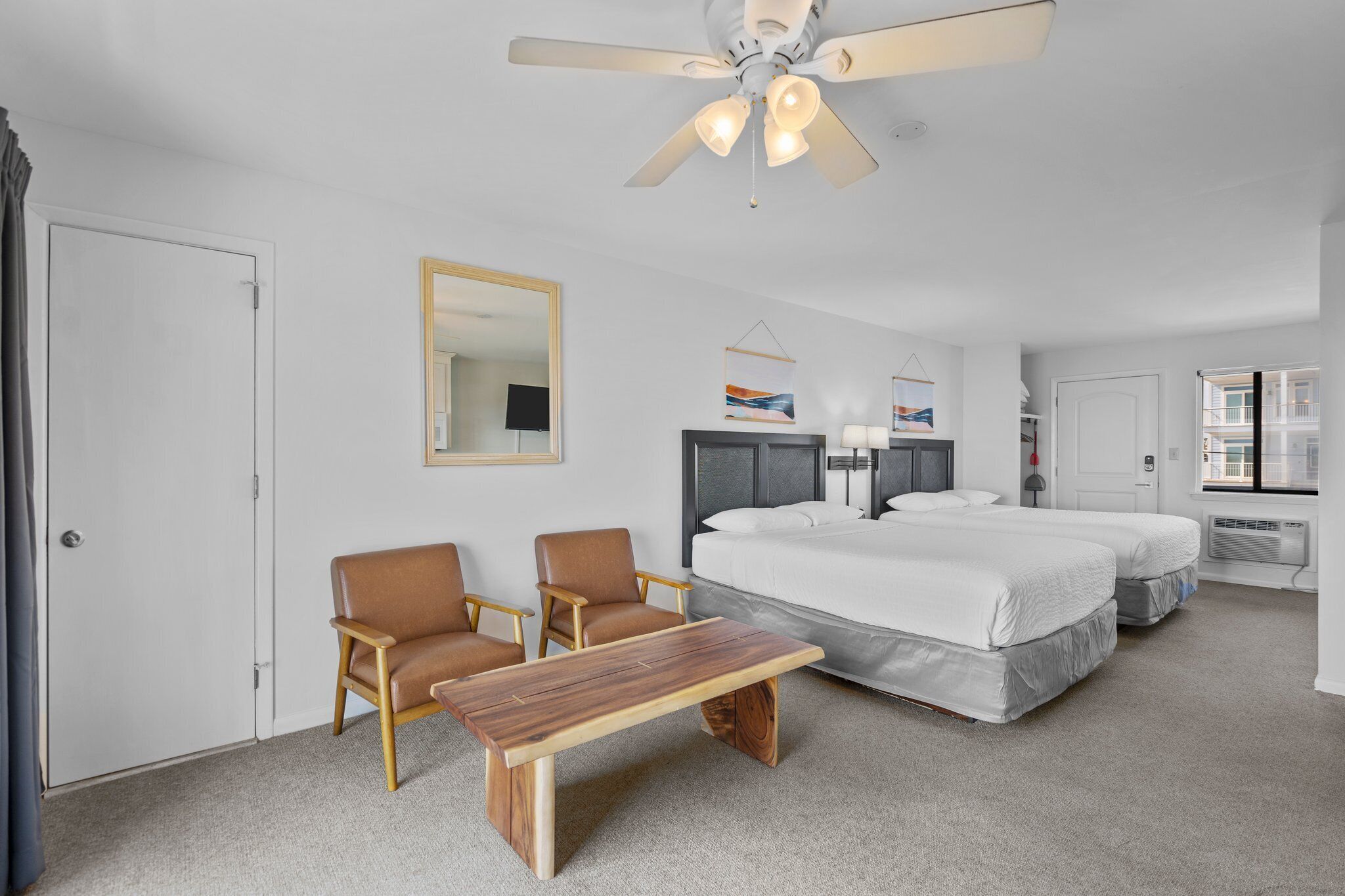 A hotel room with two beds, a wooden table, two armchairs, a ceiling fan, and a large mirror.