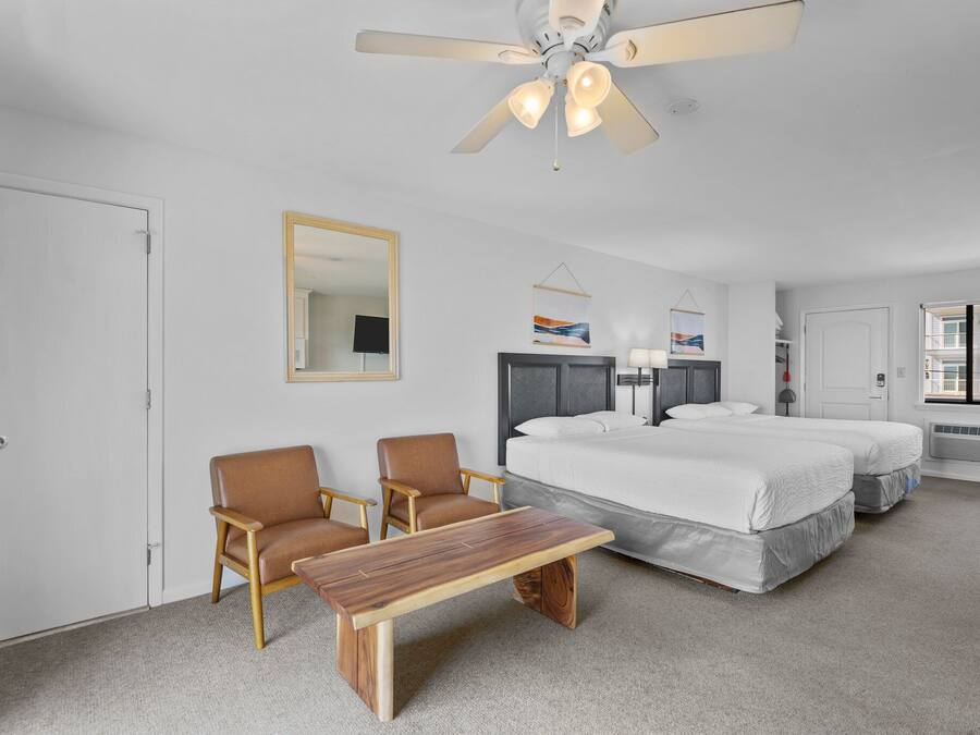 Ein Hotelzimmer mit zwei Betten, einem Holztisch, zwei Sesseln, einem Deckenventilator und einem großen Spiegel.