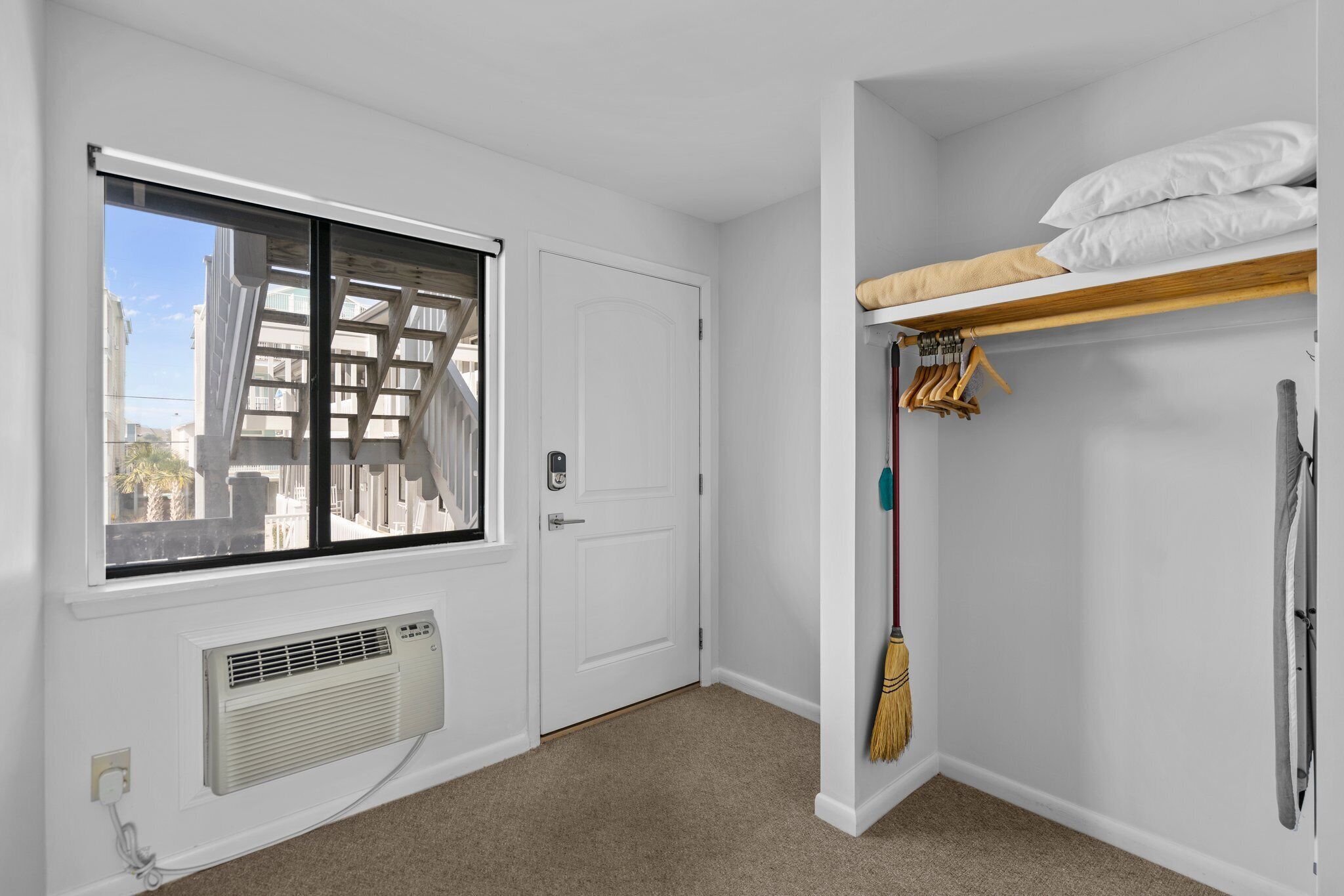 Una habitación con ventana, puerta, radiador, una cama litera con almohadas, una escoba y un perchero.