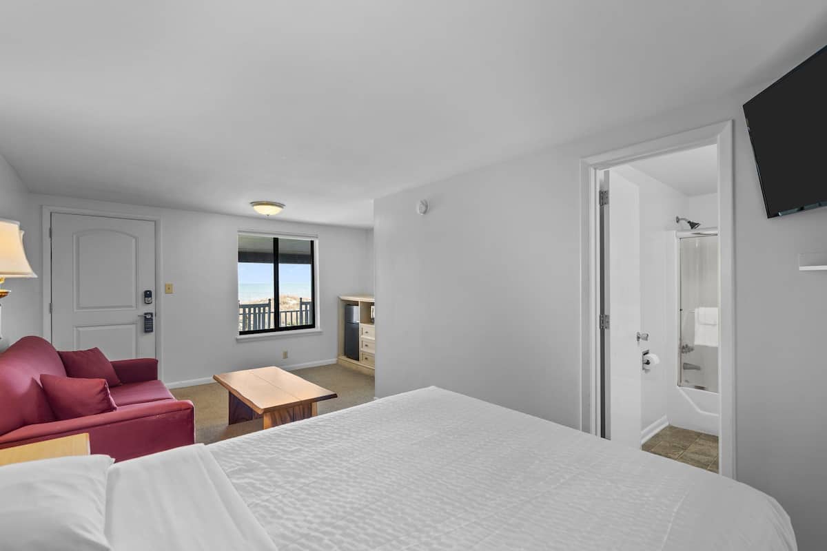 A hotel room with a bed, a red sofa, a small table, a desk, a TV, a window with a view, and a bathroom with a bathtub.
