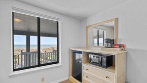 A room with a large window overlooking a beach, a kitchenette with a refrigerator, microwave, and coffee maker, and a wooden cabinet with drawers. - The Beach House (Carolina Beach)