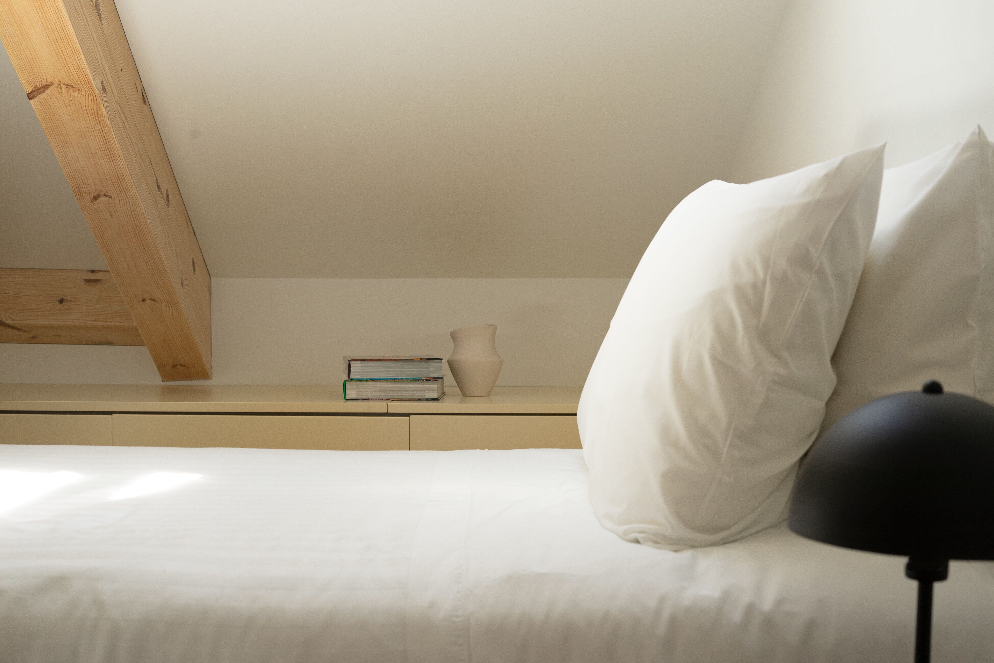 Una camera da letto con un letto, cuscini, un comodino con una lampada, un libro e un vaso.