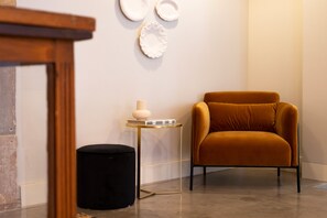 A room with a mustard-colored chair, a small round table with a white lamp, and a black ottoman. - Entre Ruas Apartments (Coimbra)