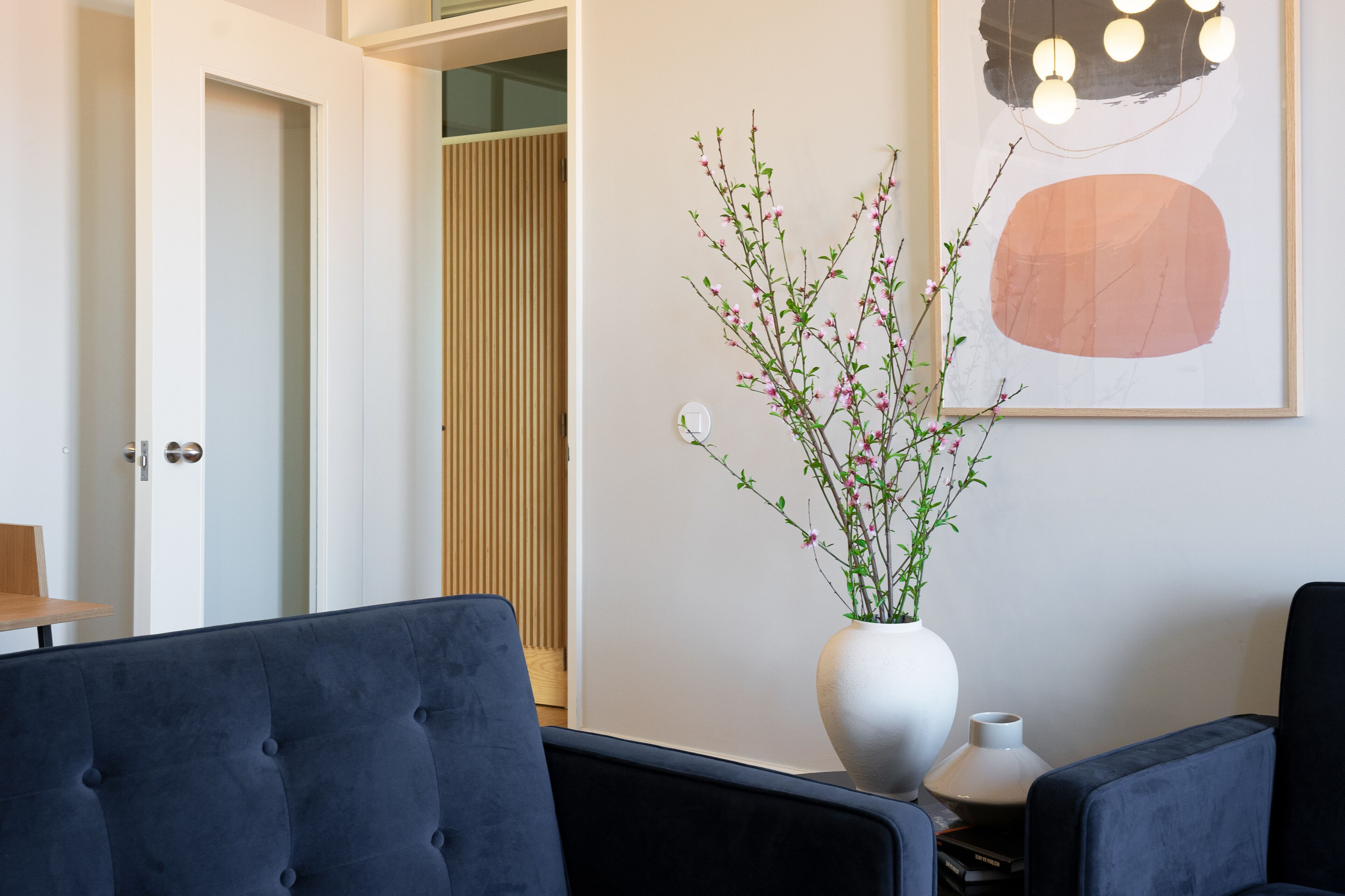 A modern room with a dark blue sofa, a white vase with pink flowers, and a framed abstract painting.
