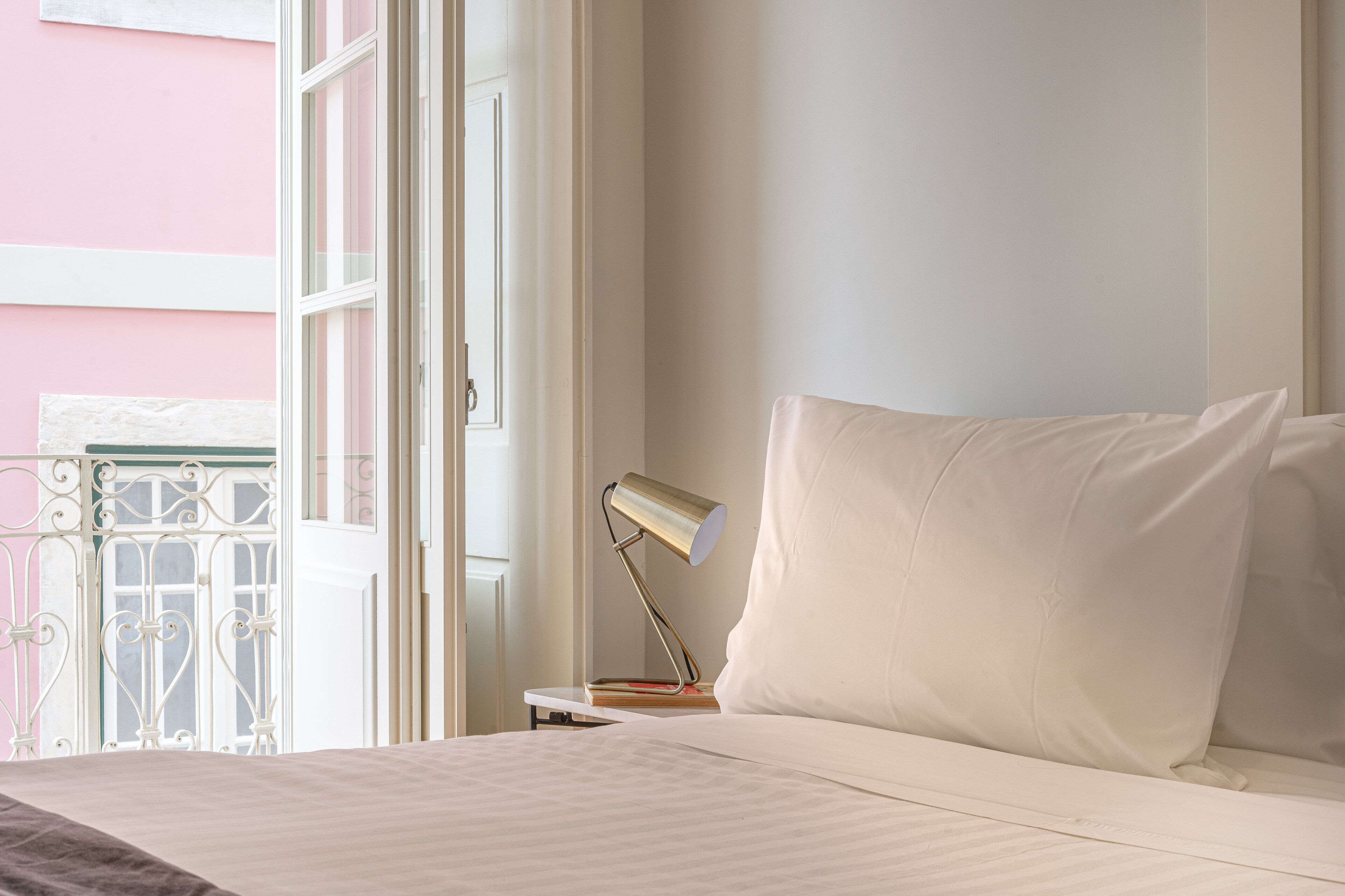 A neatly made bed with white linens, a bedside table with a lamp, and a view of a pink building through a window.