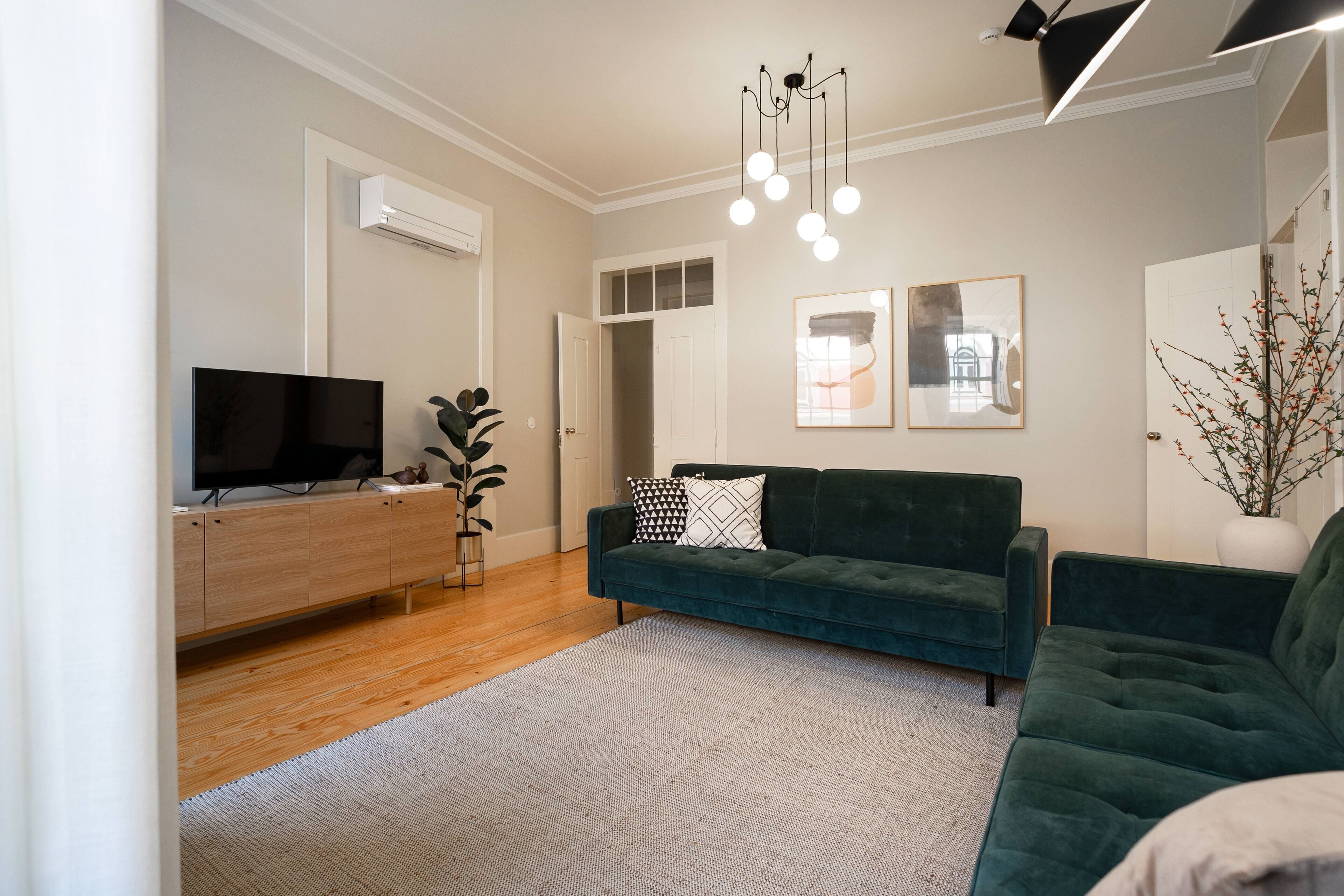 Ett modernt vardagsrum med en mörkgrön soffa, en platt-TV placerad på ett träskåp, en vit matta och en taklampa.