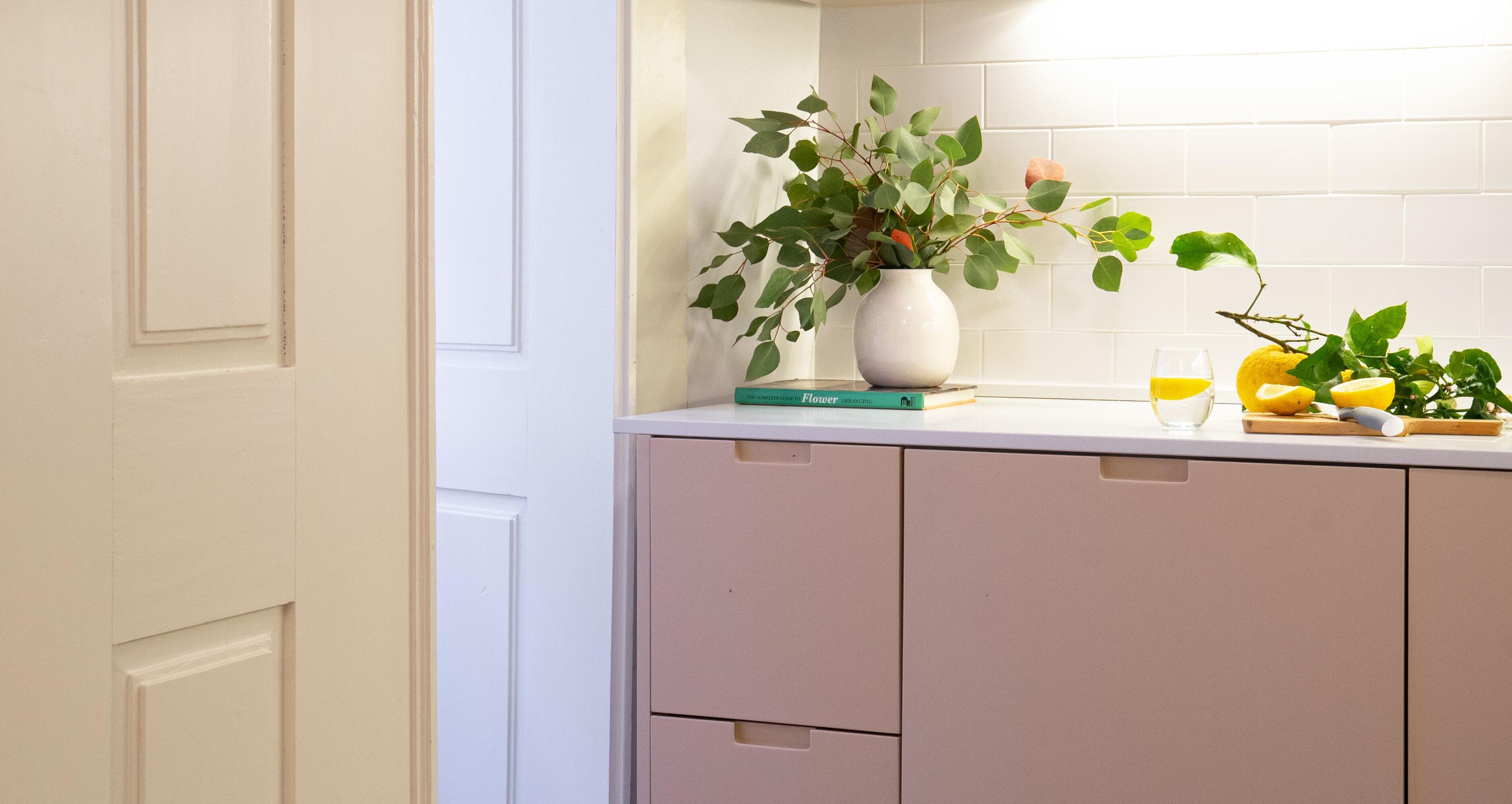 A kitchen with a white cabinet, a white vase with green leaves, a book, a glass of orange juice, and a lemon on the counter.
