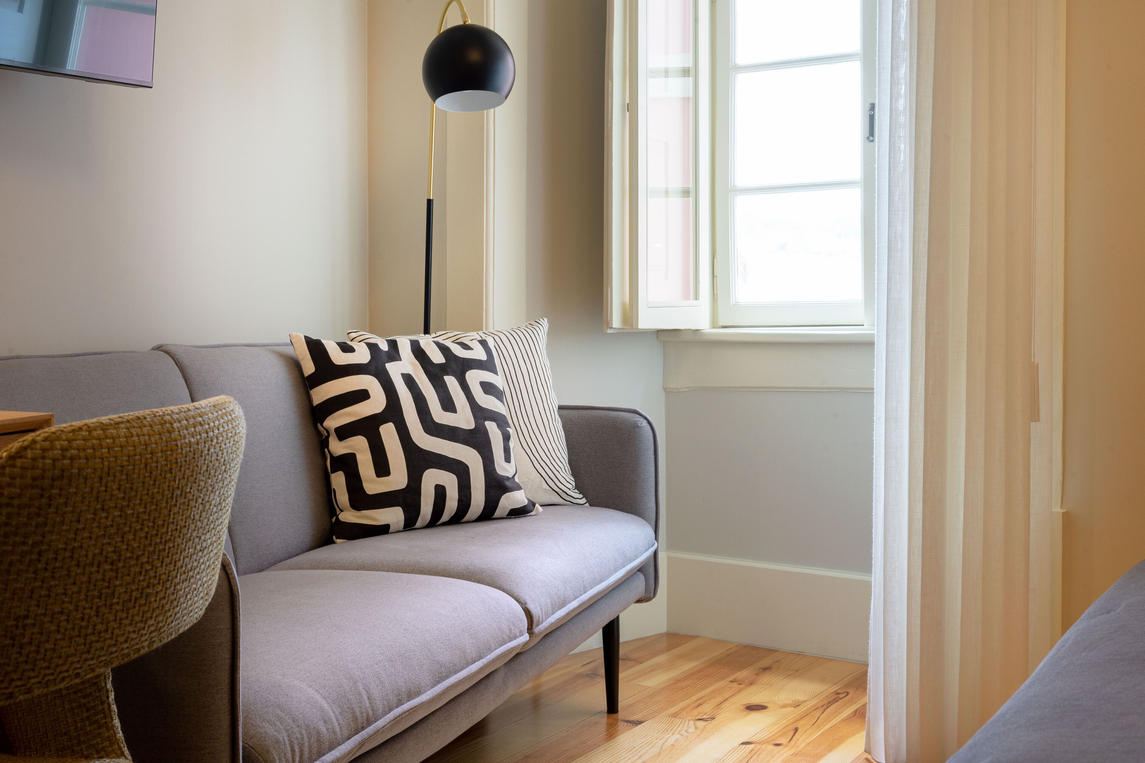 Ein modernes Wohnzimmer mit einem grauen Sofa, einem gemusterten Zierkissen, einer Stehlampe und einem Fenster mit Vorhängen.