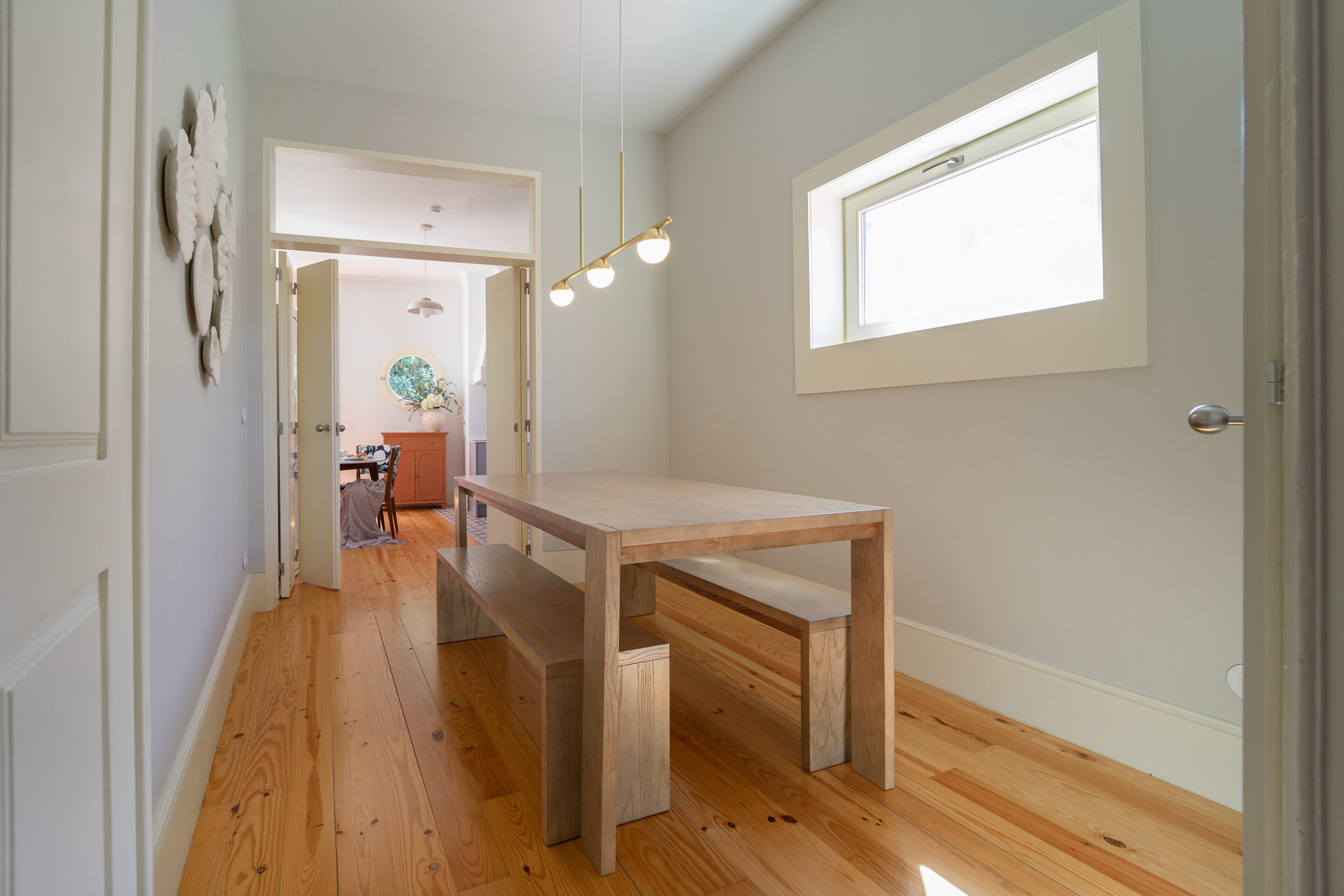 Un couloir avec une table et un banc en bois, une fenêtre avec des stores et une porte donnant sur une autre pièce.