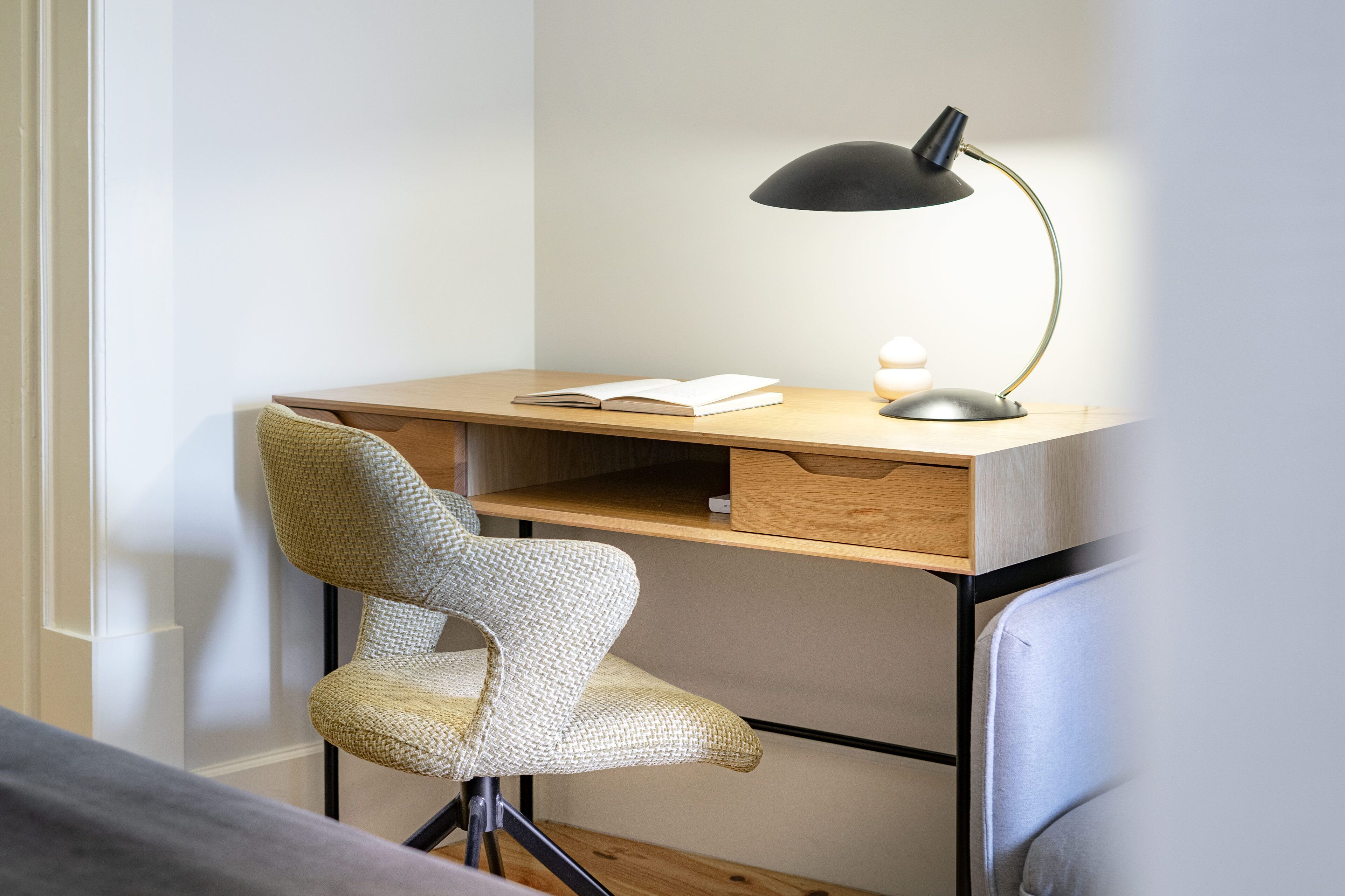 En modern arbetsplats med ett skrivbord i trä, en svart skrivbordslampa, en flätad stol och ett litet vitt ljus.