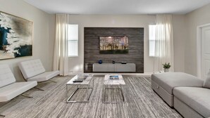 Un salon moderne avec un canapé gris, deux tables basses en marbre, un canapé d’angle gris, un tapis gris à motifs, un mur gris avec un panneau en bois, une grande peinture abstraite, un téléviseur à écran plat et une petite plante.