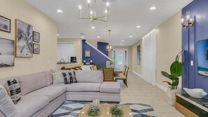 A modern living room with a large L-shaped sofa, a coffee table with plants, and a dining area with a staircase in the background. - Last Minute Disney! Private Pool, Hot Tub, Game Room & Themed Bedrooms. Book Now! #65729 (Orlando)