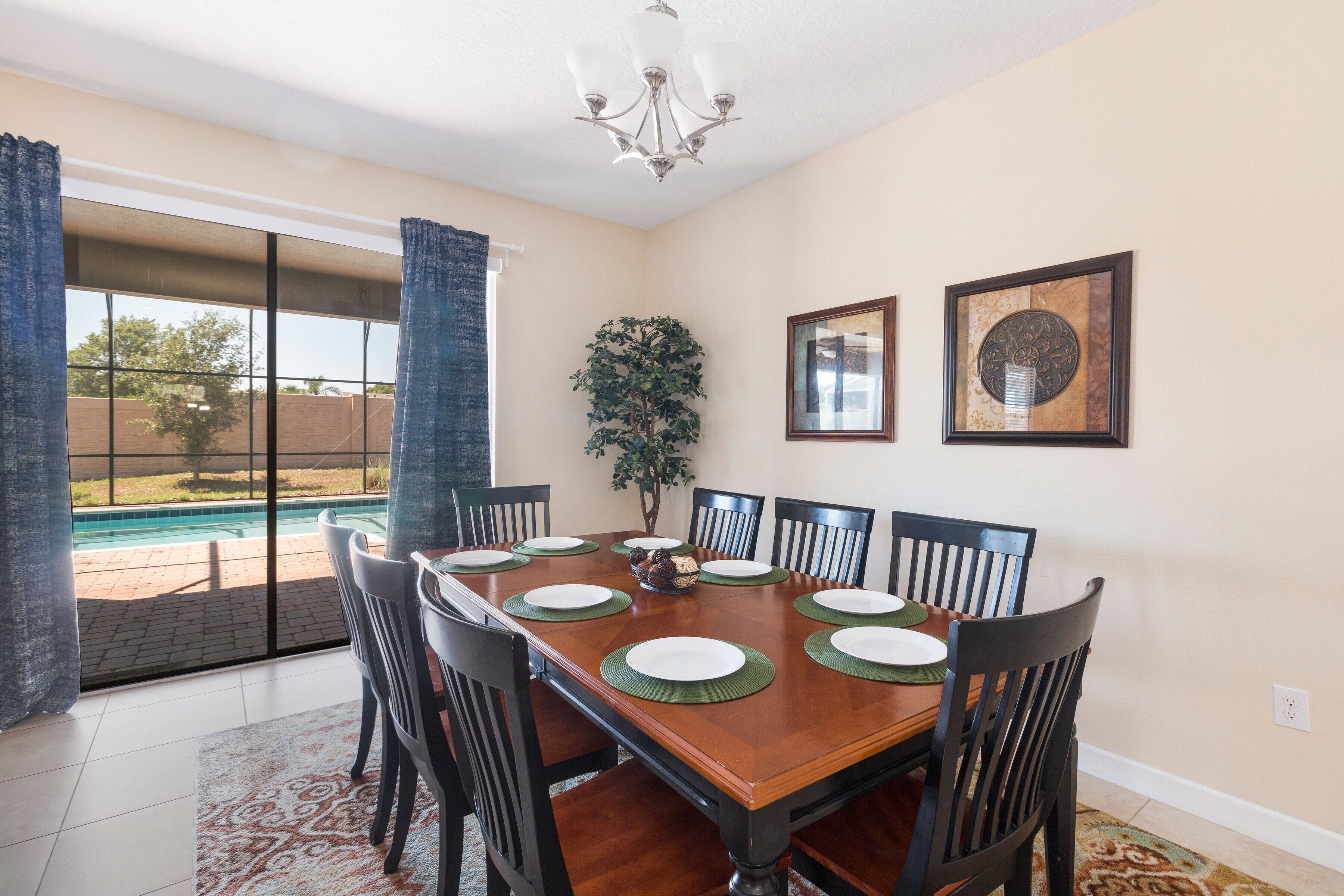 Comedor con una mesa de madera dispuesta para ocho personas, rodeada de sillas, un candelabro y un gran ventanal con cortinas.