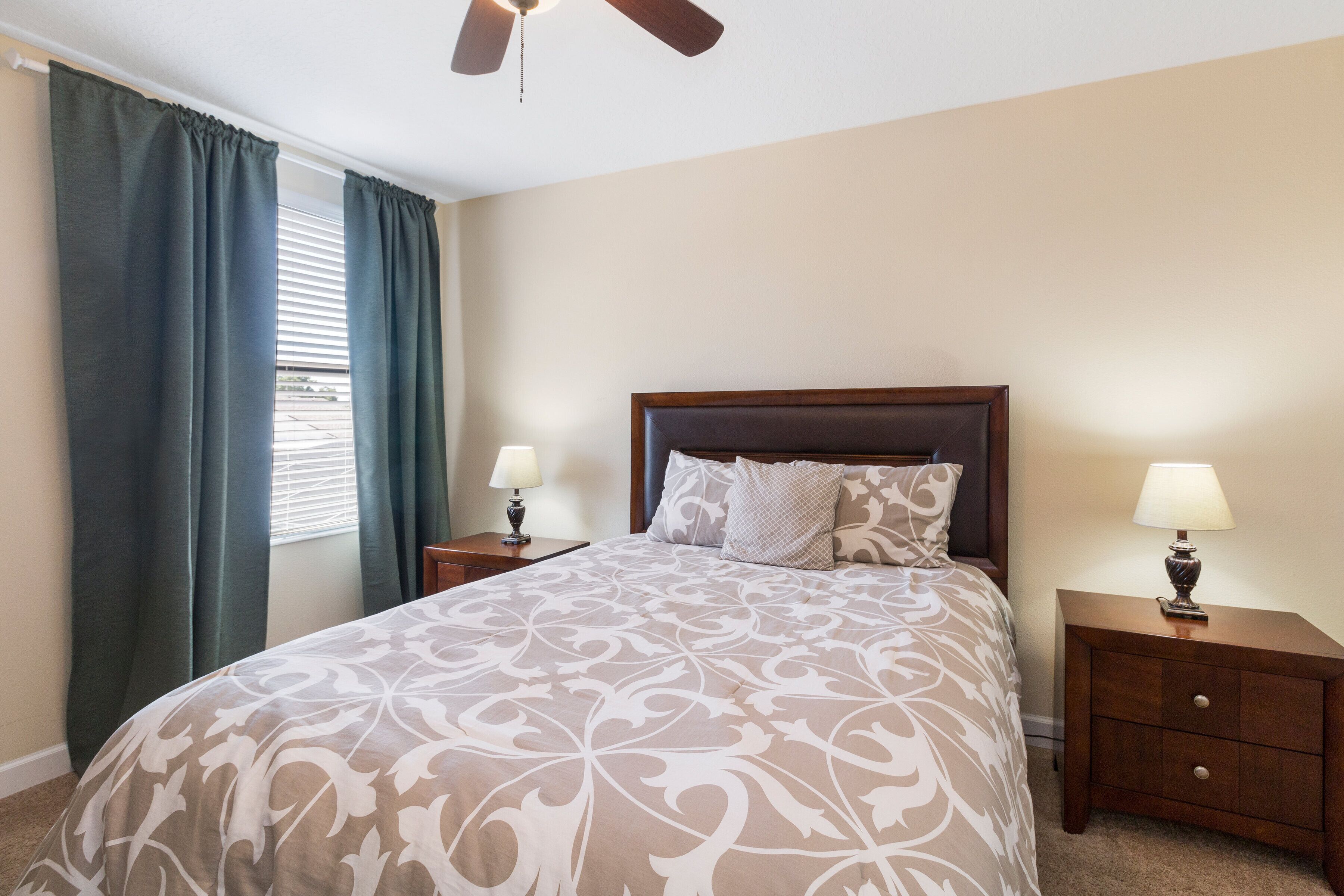 A bedroom with a large bed, two bedside tables with lamps, a ceiling fan, and a window with curtains.
