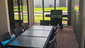 Un patio techado con una mesa y sillas, un ventilador de techo y vistas a un jardín trasero con una parrilla.