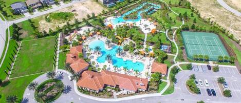 An aerial view of a resort with a central pool, tennis courts, and a parking area.