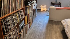 Una habitación con una amplia estantería llena de libros y DVD, una cama, una mesita con lámpara y un ventanal con persianas.