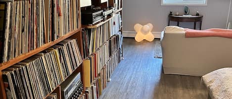 A room with a large bookshelf filled with books and DVDs, a bed, a small table with a lamp, and a window with blinds.