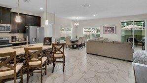 A modern kitchen with dark cabinets, a central island with chairs, and a dining area with a table and chairs. - Last Minute Disney Getaway! Private Pool & Themed Bedrooms. Book Now! #65717 (Orlando)