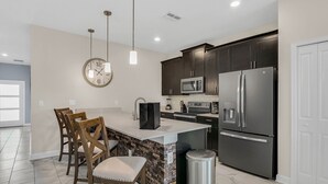 A modern kitchen with dark cabinets, stainless steel appliances, a large island with seating, and a clock on the wall. - Last Minute Disney Getaway! Private Pool & Themed Bedrooms. Book Now! #65717 (Orlando)