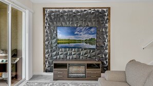 A living room with a flat-screen TV mounted on a geometric-patterned wall, a wooden entertainment unit below, and a beige sofa. - Last Minute Disney Getaway! Private Pool & Themed Bedrooms. Book Now! #65717 (Orlando)