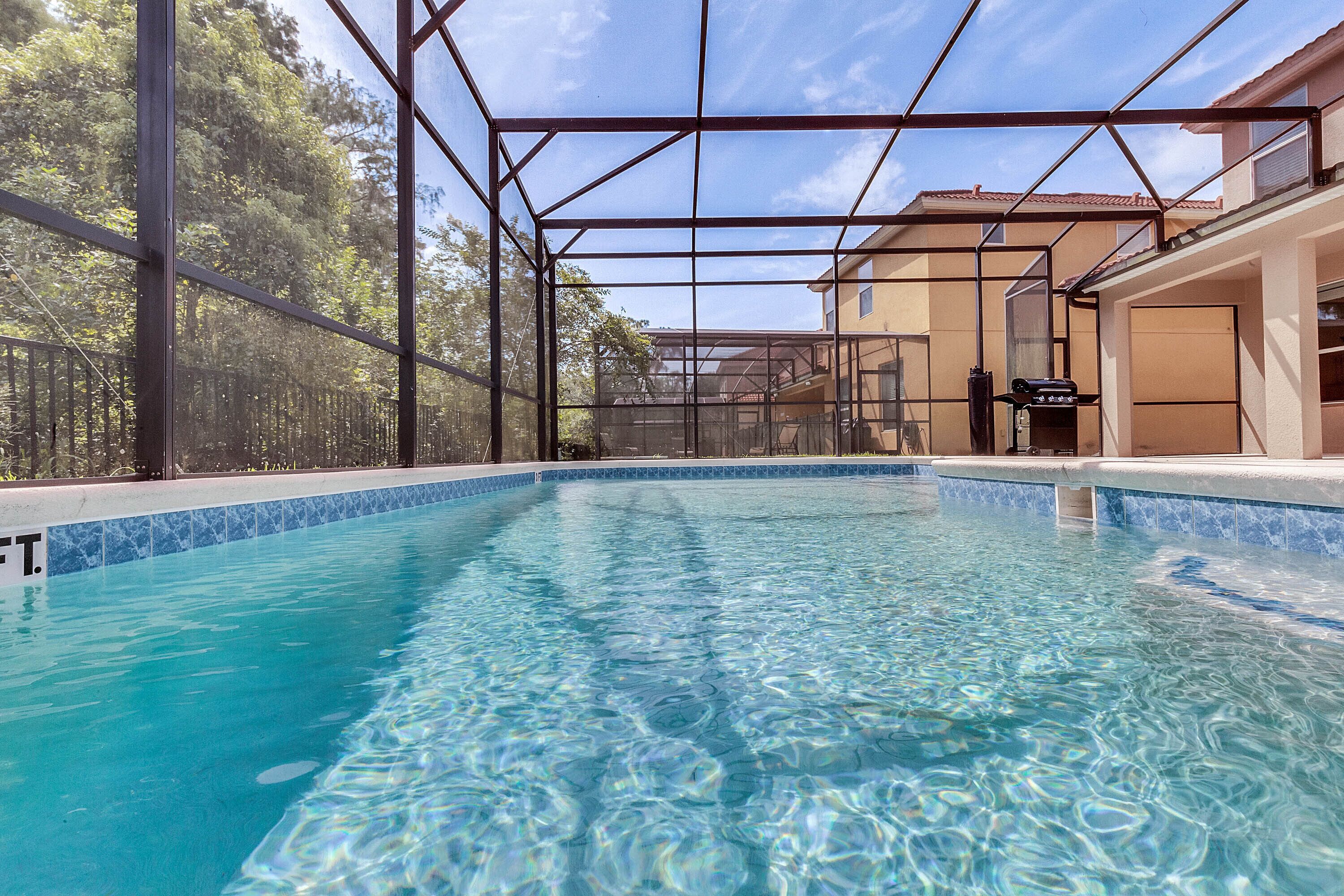 A clear indoor swimming pool with a glass ceiling, surrounded by a metal frame, and a view of a building with a tiled roof in the background.