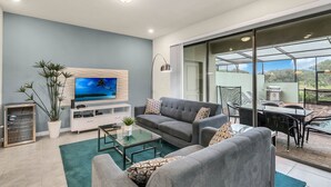 A modern living room with a grey sofa, a flat-screen TV, a glass coffee table, and a dining area with a table and chairs.