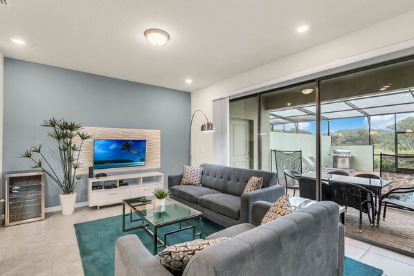 A modern living room with a grey sofa, a flat-screen TV, a glass coffee table, and a dining area with a table and chairs.