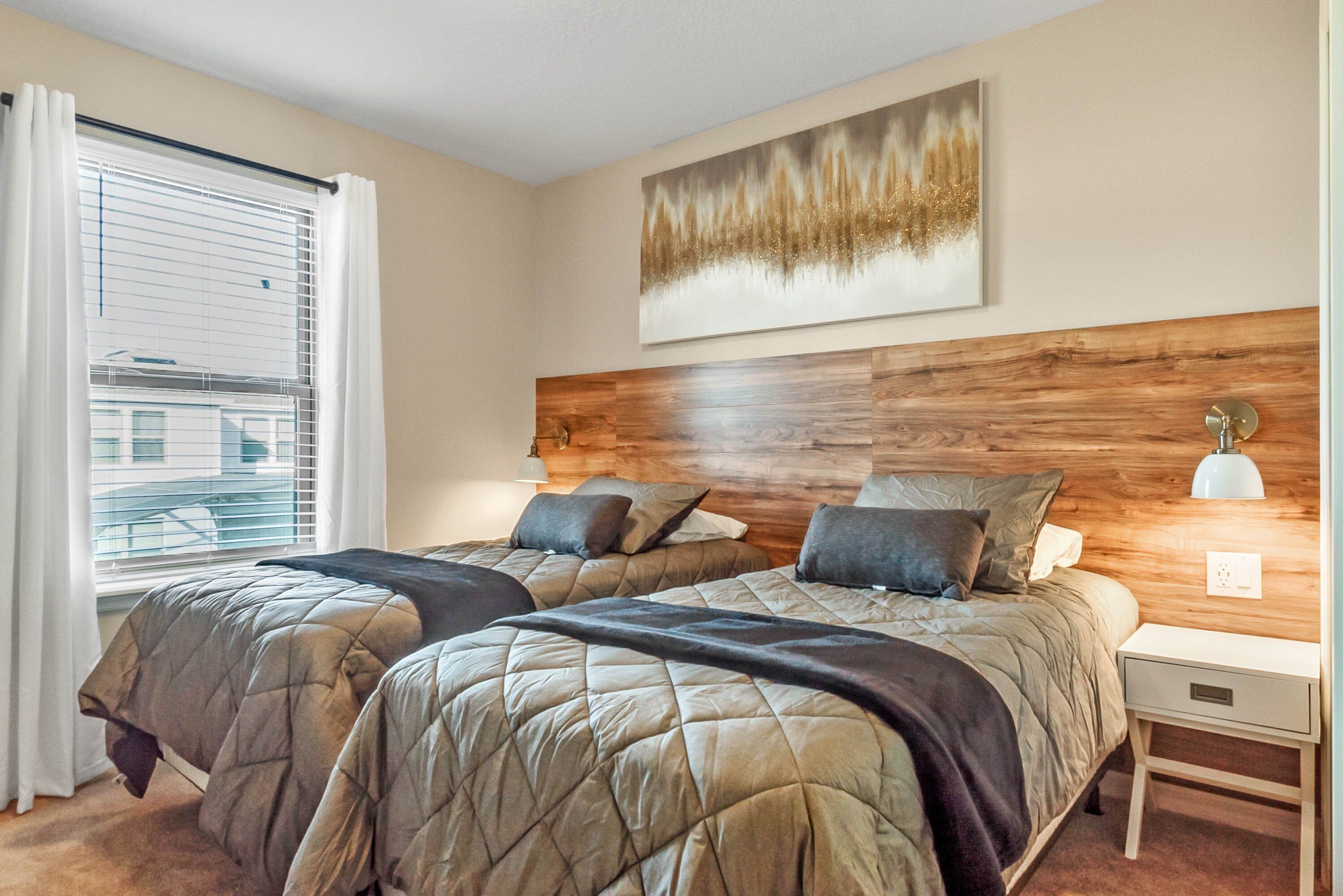 A hotel room with two beds, a wooden headboard, a painting, a bedside lamp, and a window with curtains.