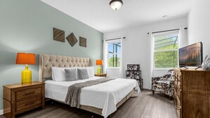 Un dormitorio con cabecera capitonada, dos mesitas de noche con lámparas, cama con colcha blanca, cómoda de madera y una ventana grande con vistas a la vegetación.