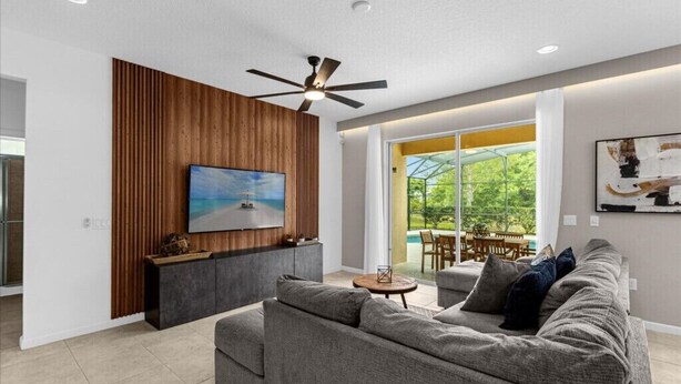Una sala de estar moderna con un sofá gris, un televisor de pantalla plana montado en una pared de paneles de madera, un ventilador de techo y vistas a un patio con mobiliario exterior.