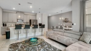 A modern kitchen with bar stools, a dining area with a grey sofa, and a chandelier. - Last Minute Disney Getaway! Private Pool & Themed Bedrooms. Book Now! #65827 (Orlando)