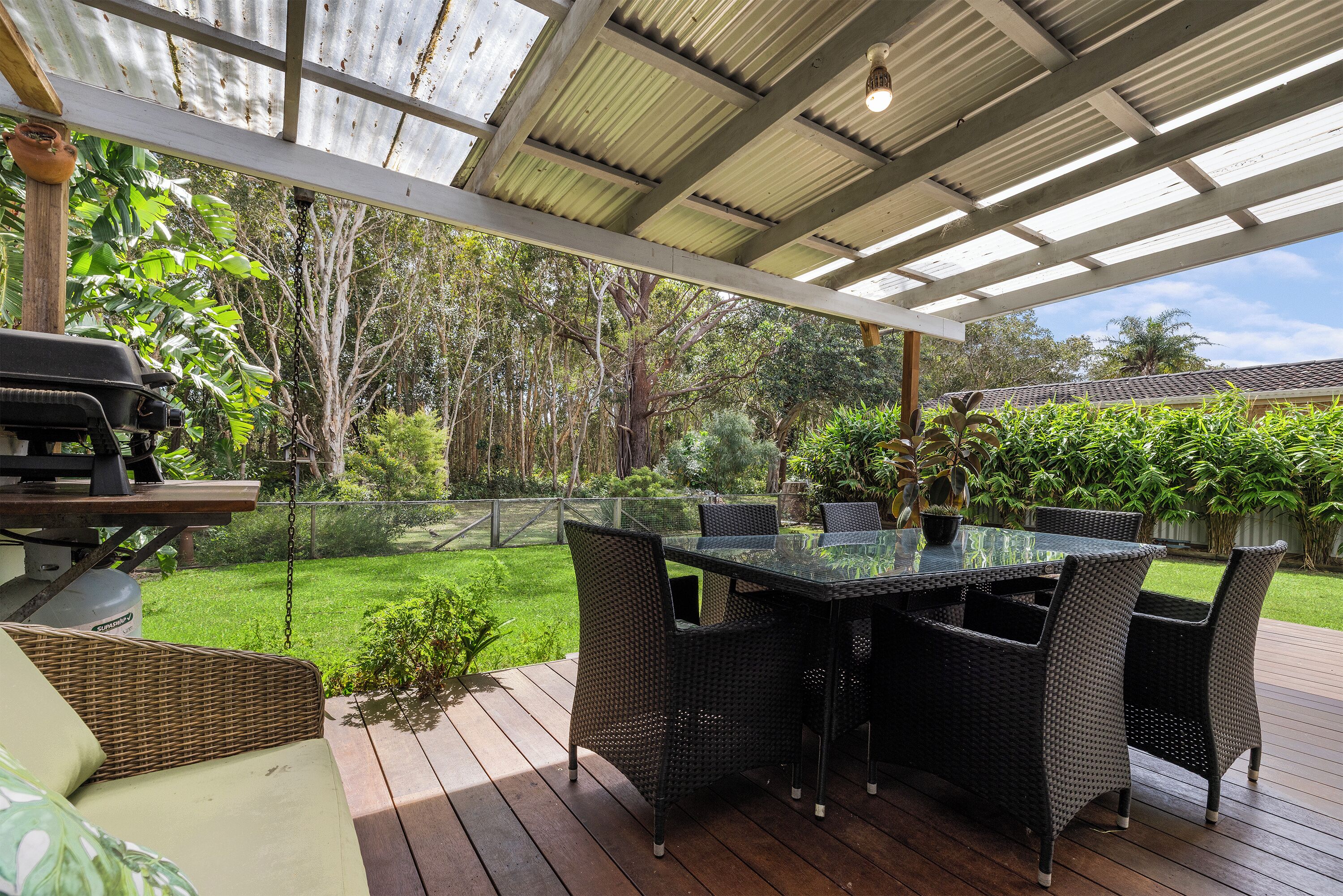 Une terrasse extérieure dotée d’une table à plateau de verre et de chaises en osier, entourée d’une plateforme en bois et offrant une vue sur un jardin luxuriant.