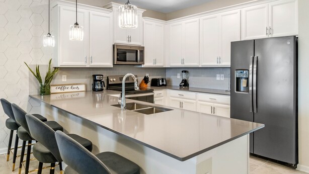 A modern kitchen with white cabinets, a stainless steel refrigerator, a central island with bar stools, and pendant lighting.
