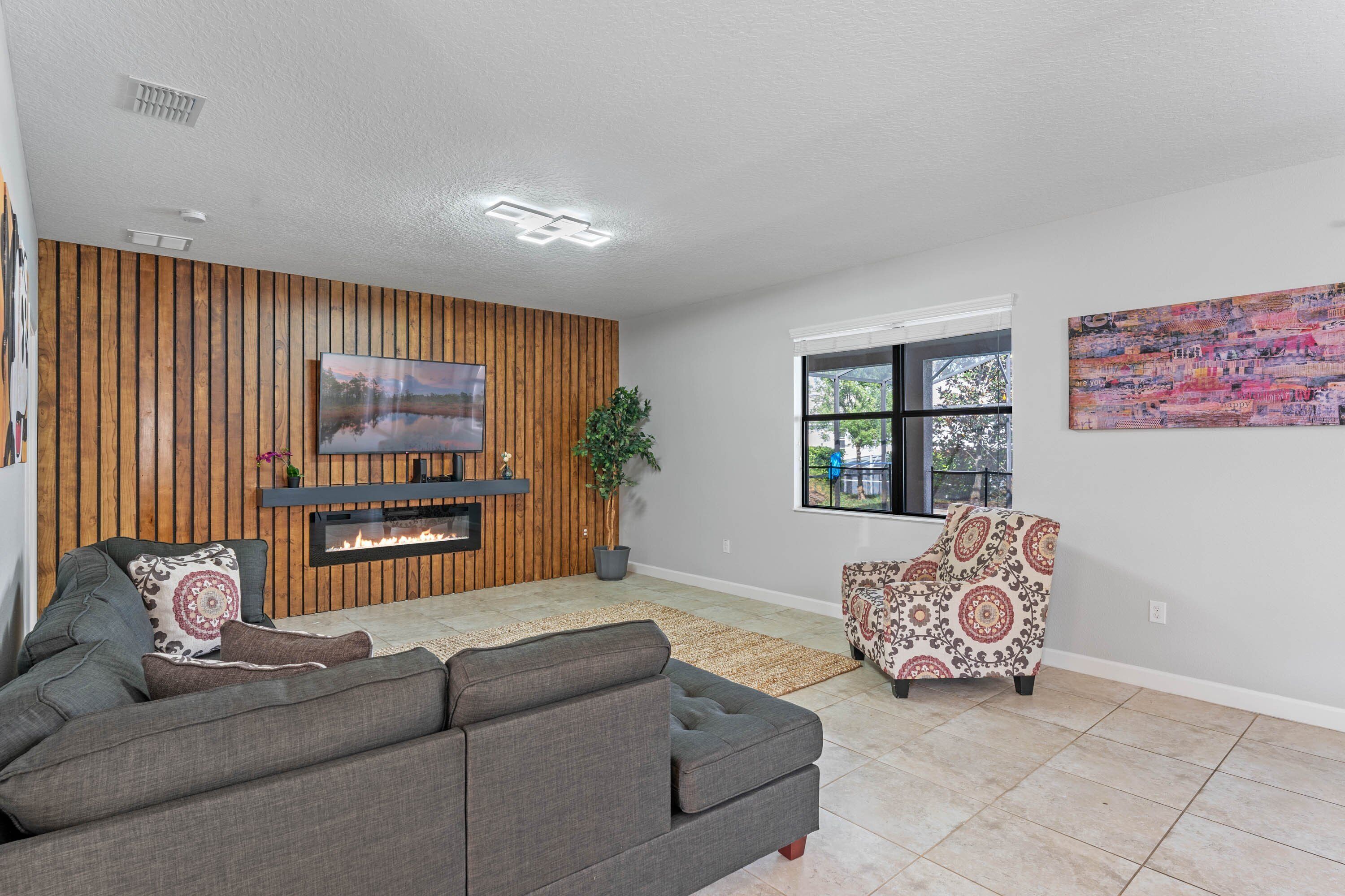 A living room with a gray sectional sofa, a patterned armchair, a wooden wall panel, a fireplace, and a large window with a view.