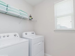 A laundry room with two white washers and dryers, a shelf with folded towels, and a window with blinds.