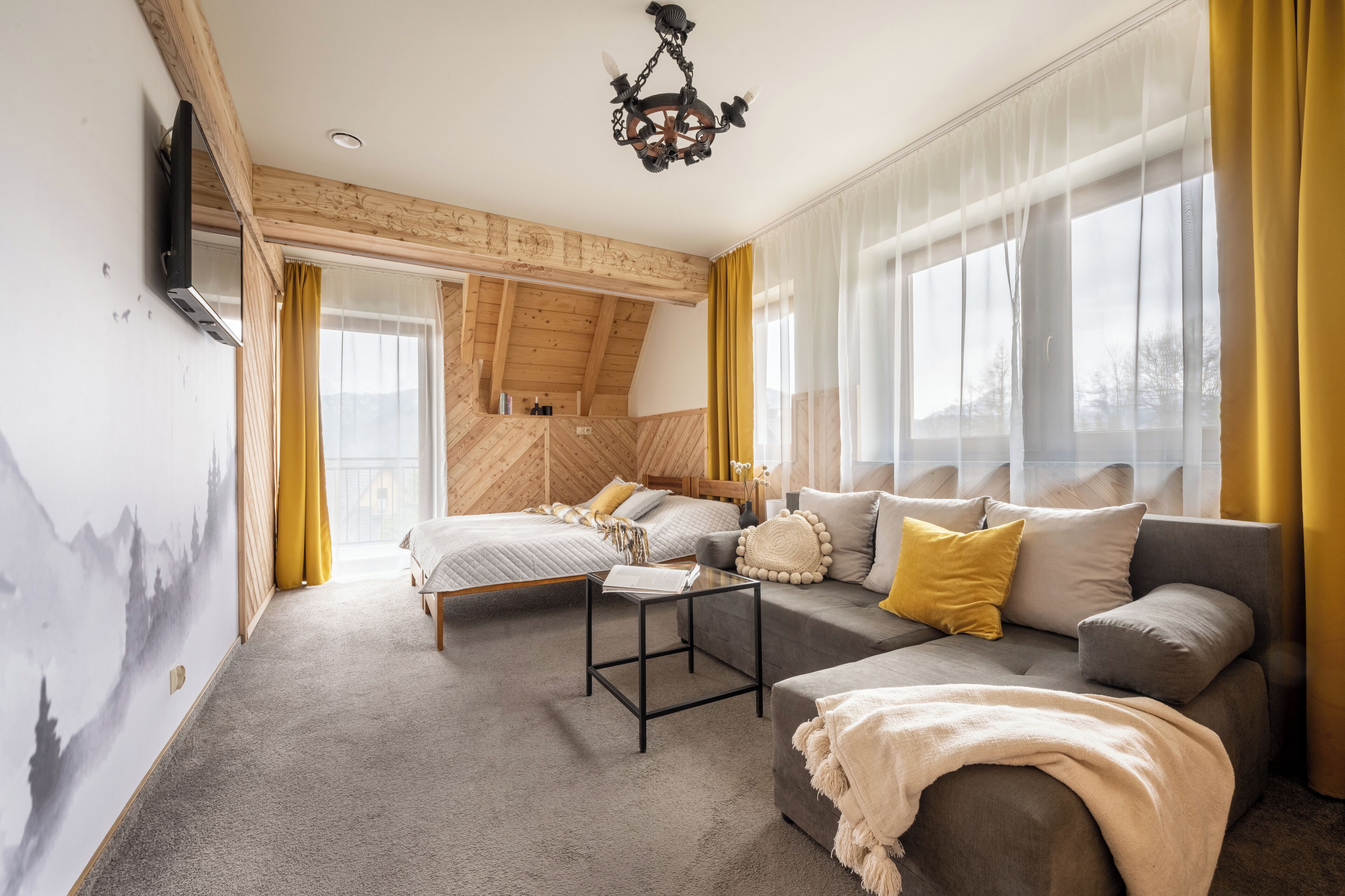 A cozy room with a bed, sofa, and a small table. There is a window with curtains, a ceiling light, and a wall-mounted TV.