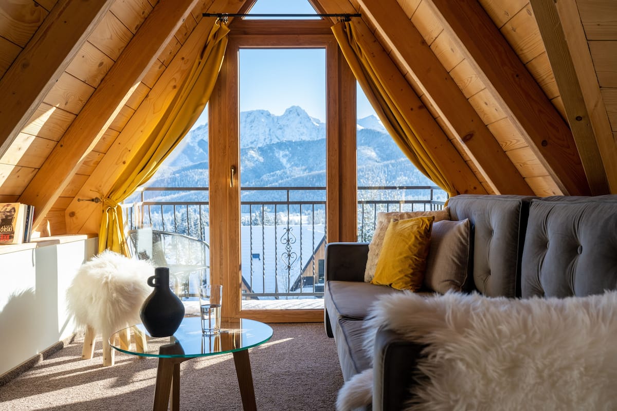 Une pièce chaleureuse avec vue sur des montagnes enneigées, une table en verre, un canapé et un chien blanc et duveteux.