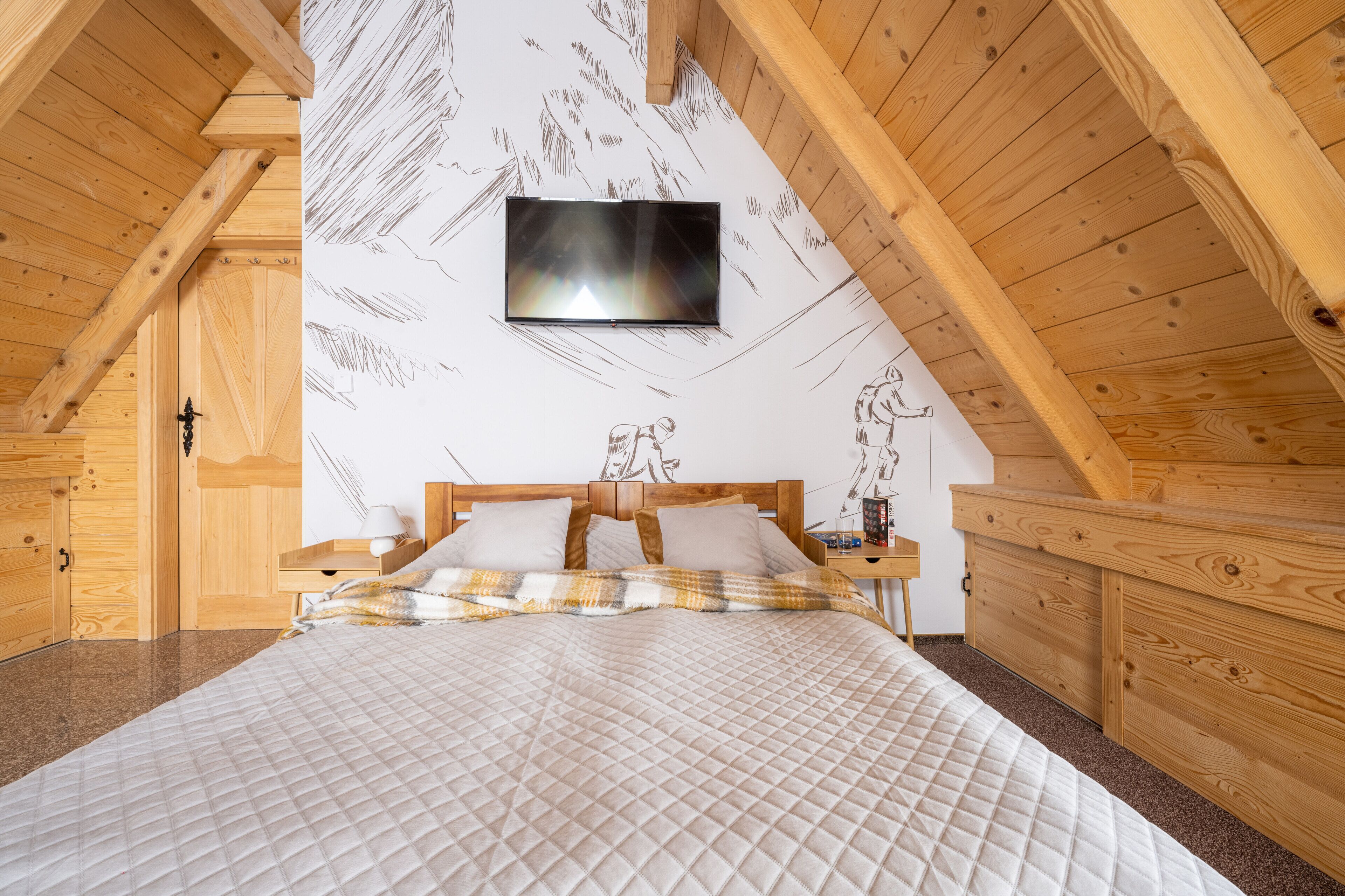 A bedroom with a wooden ceiling, a bed with a quilted bedspread, a mounted TV, and a nightstand with a lamp.
