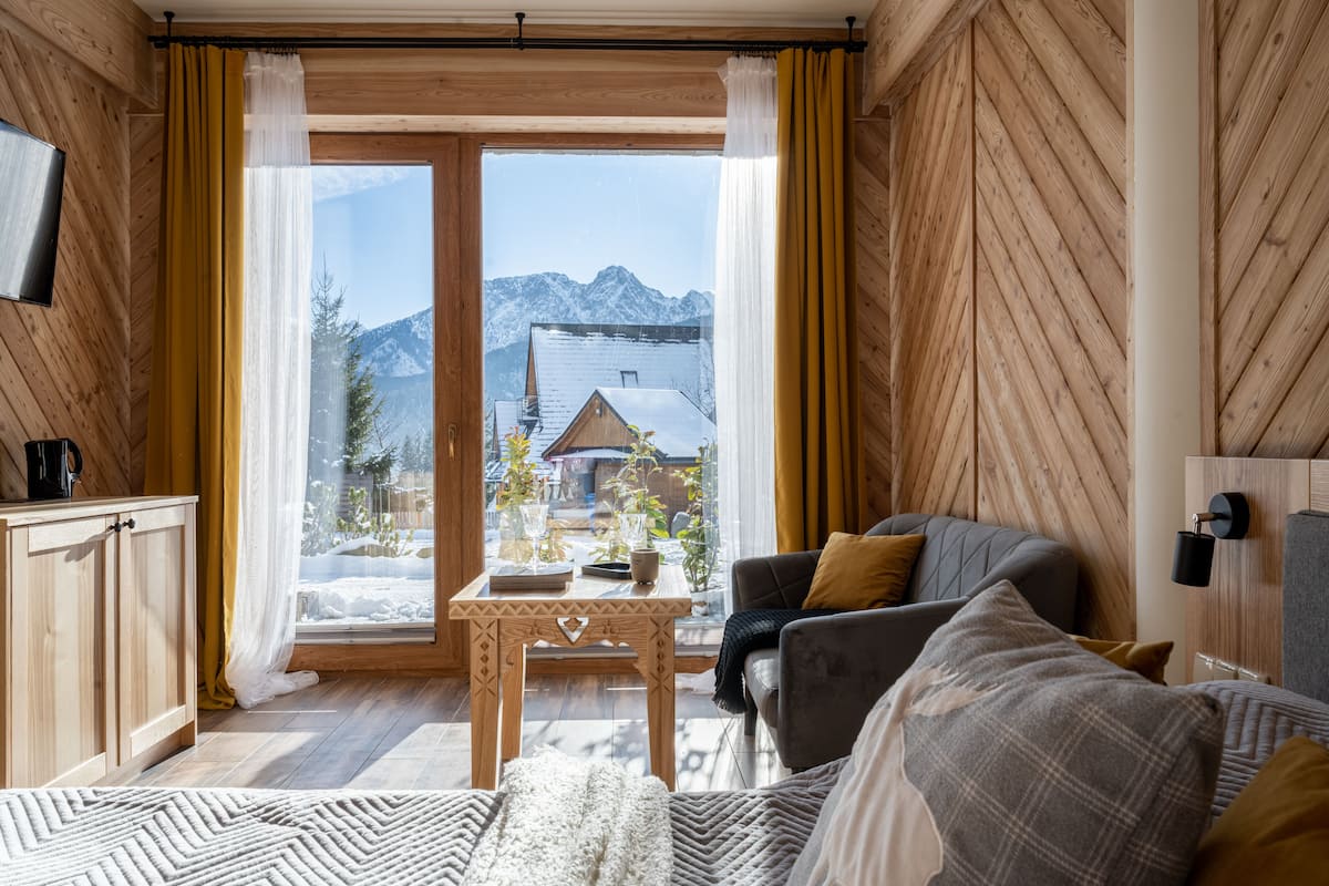 Une pièce chaleureuse aux murs en bois, avec un canapé, une table et une vue sur les montagnes par une grande fenêtre.